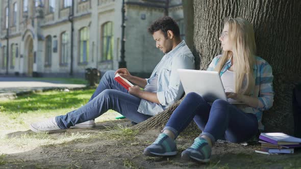 Guy Reading Book Under Tree, Girl Using Laptop Casting Looks at Each Other