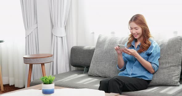 Asian woman relaxing on the couch and browsing social media on her smartphone. Happy girl texting he