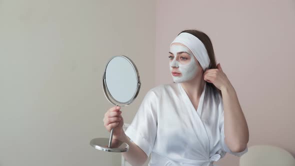 Young Woman in Beauty Cosmetology Salon with Clay Mask on Face Looks at the Mirror