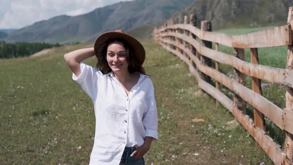 A Young Woman in Shorts and a White Shirt Walks Along a Fence in Rural locality.Attractive Brunette