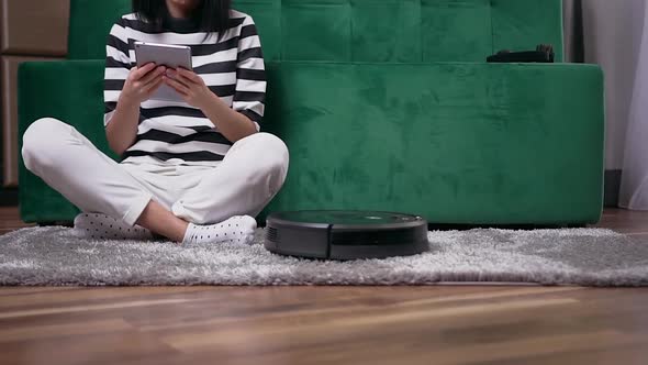 Woman Sitting on the Carpet Near Green couch while Robotic Vacuum Cleaner Cleaning the Room