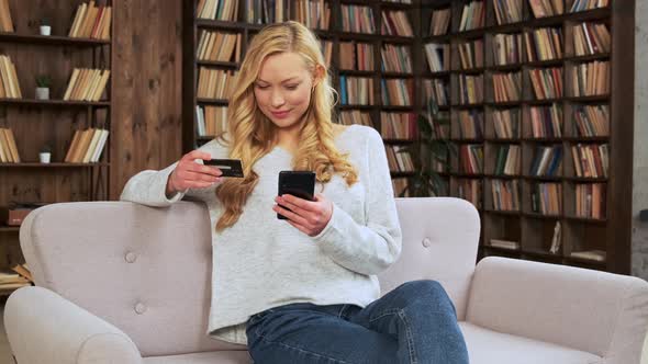 Young Blonde Woman Sitting on the Couch and Makes Purchases Online Using Credit Card and Smart Phone