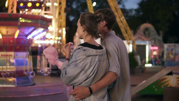 Backside View of a Young Dating Couple Walking By Funfair at Night Eating Cotton Candy