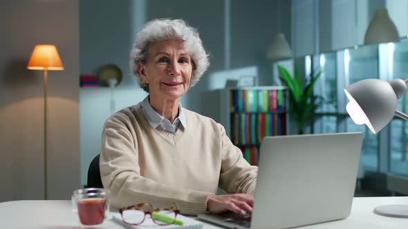 Portrait of Happy Aged Woman Smiling at Camera Sitting with Laptop at Desk in Living Room