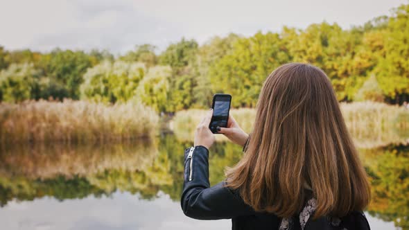 Young Woman with Backpack Taking Photos of Autumn Landscape Capturing Lake and Forest Nature on