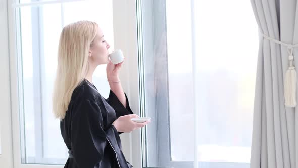 Young Beautiful Woman in Silk Black Robe Drinking Coffee Near the Window at Home