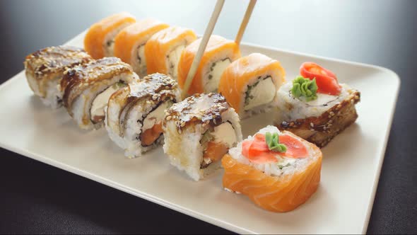 Hand Holds Bamboo Sticks and Shifts Sushi on a White Plate.