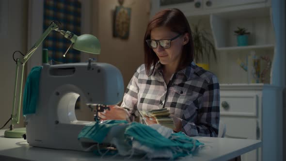 Happy 30s woman counting dollars money for sewing protective face mask working from home