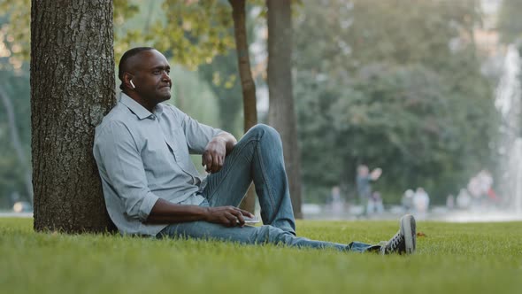 Happy Elderly African Black Businessman Wearing Wireless Headphones Sitting on Grass in City Park