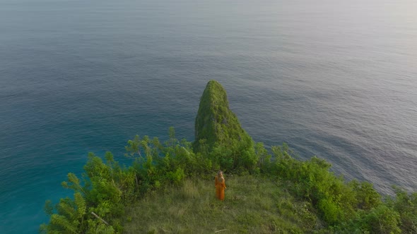 Woman in orange dress looking in awe at amazing tropical green cliff during sunset, Sekartaji cliff