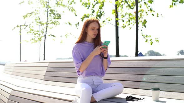 Happy Hipster Redhead Girl Communicating By Mobile Phone Outdoors While Sitting on Bench in Park.