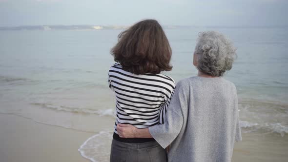 Rear View of Happy Senior Woman Embracing Tall Brunette on Seashore