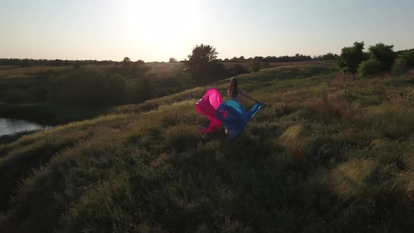 A Girl with Loose Hair in a Dress Plays with a Pink and Blue Cloth on the High Bank of the Lake