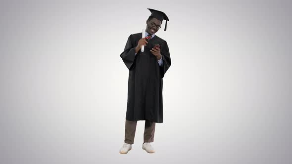 Smiling African American Male Student in Graduation Robe with Diploma Texting on the Phone on