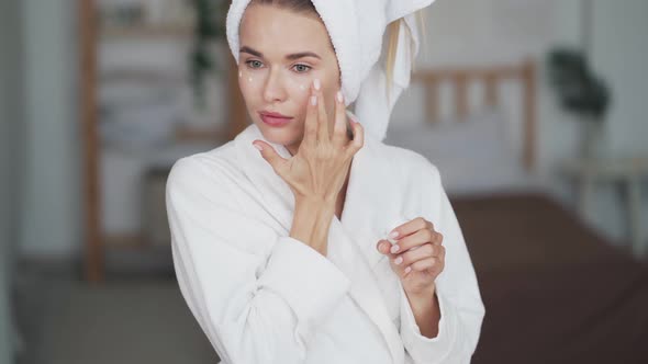  Woman with Towel on Head Applies Moisturizer Cream on Her Face, Close Up