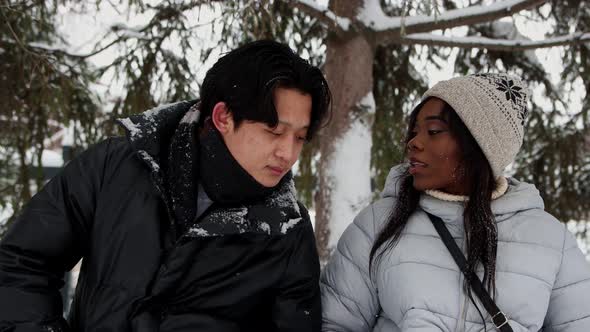 Young Asian Man Talking with His Black Woman Friend Sitting on Bench in Winter Park