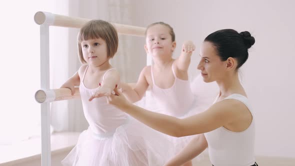Two Little Girls Practicing Choreographic Elements on Ballet Barre with Help of Teacher in Dance