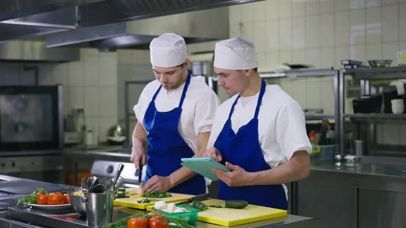 Caucasian Male Cook Reading Recipe on Tablet Consulting Professional Chef in Commercial Kitchen