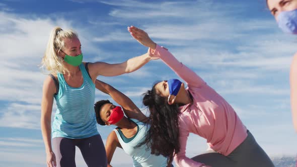 Group of diverse female friends wearing face masks practicing yoga at the beach