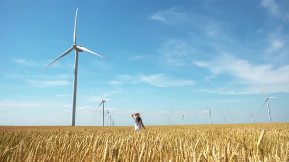 Beautiful girl walking on yellow field of wheat with windmills for electric power production