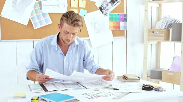 Young Successful Architector Sorting Out Drawings Writing Sitting at Workplace