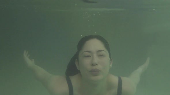 Woman swimming underwater in pool and then emerging to smile at camera