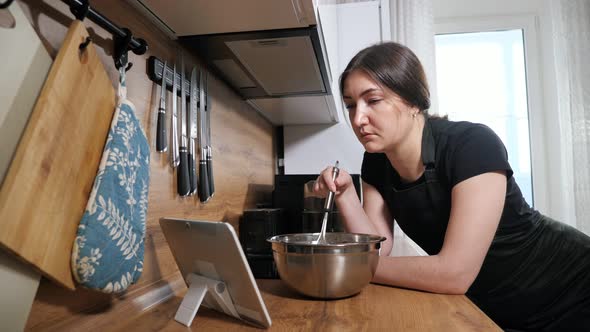 Brunette Woman Mixes with a Whisk and Looks Into a Tablet in Kitchen