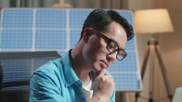 Close Up Of Asian Man Sitting In Front Of Solar Cell Looking At Wind Turbine And Thinking