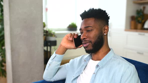 Happy Handsome AfricanAmerican Guy Has Fun Phone Conversation Sitting on the Couch at Home