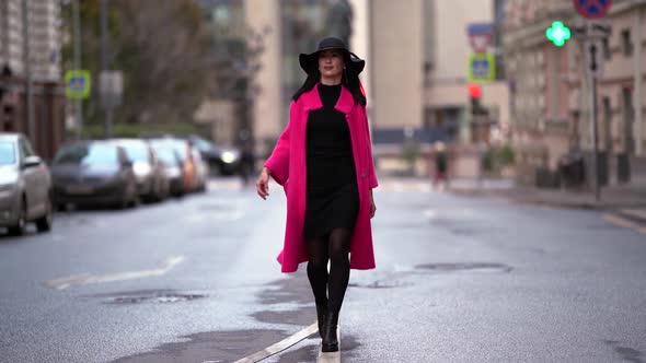 A Beautiful Brunette Woman in a Pink Coat and Black Hat Walks Along an Asphalt Road on a Blurred