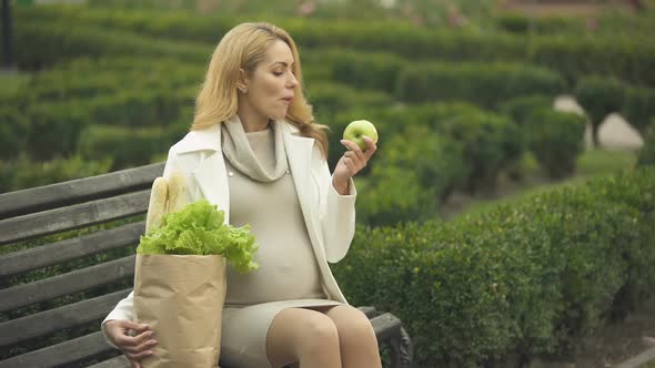 Smiling Expecting Lady Eating Fresh Apple on Bench With Grocery Bag, Health Care