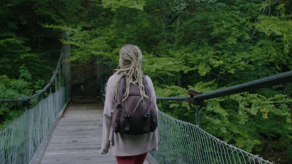 Back view Travel Woman with dreadlocks Walking Along Suspension Bridge Alone over the river