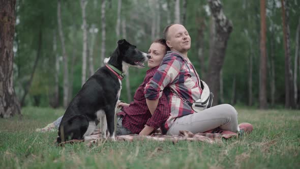 Wide Shot of Black and White Dog with Loving Couple Sitting Back To Back on Summer Lawn in Forest