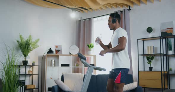 Man Listening Music in Headphones and Drink Water During Training on Treadmill at Home