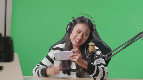 Upset Asian Woman With Headphone Losing Mobile Phone Game While Shooting Video On The Green Screen
