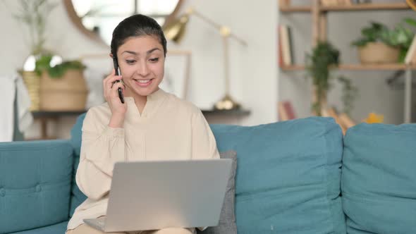 Indian Woman with Laptop Talking on Smartphone on Sofa 