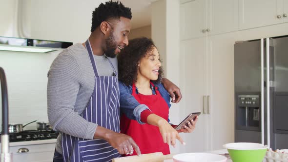 Happy african american couple wearing aprons, using tablet and baking together