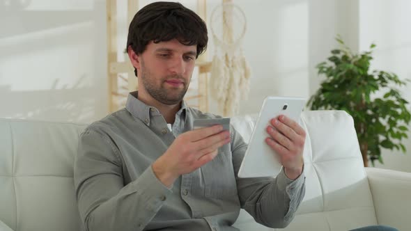 A Smiling Black Haired Man with a Tablet Computer and a Credit Card at Home Makes Purchases Online