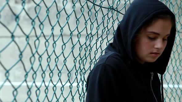 Sad schoolgirl in hooded leaning against fence