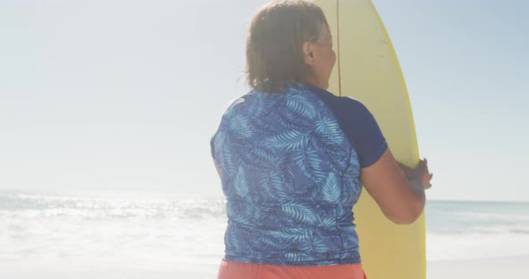 Senior african american woman standing with surfboard on sunny beach