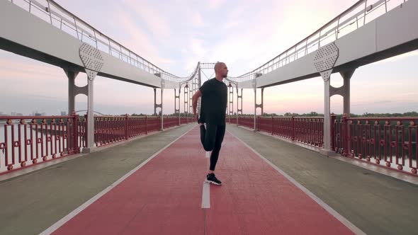 Man Runner in Black Sport Uniform Stretching Legs Before Sports on Pedestrian Bridge at Dawn