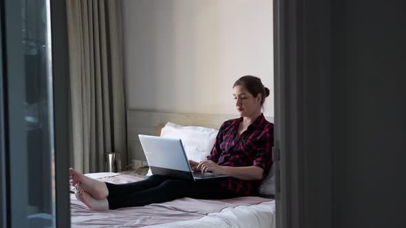 Lady Sits on White Bed and Types on Grey Modern Laptop