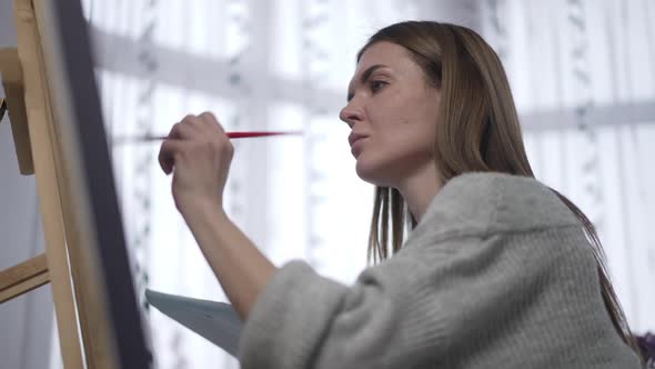 Side View Portrait of Confident Focused Woman Painting with Brush on Canvas Sitting Indoors at Easel