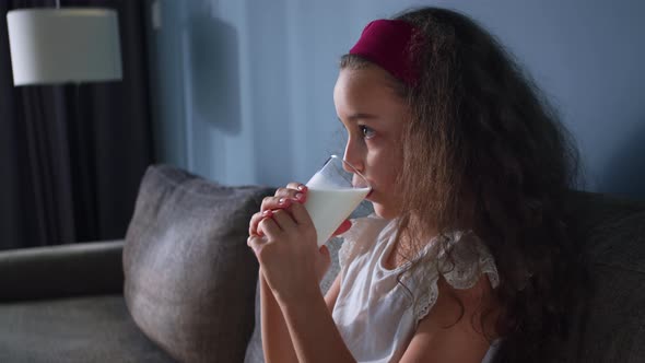 Positive Child Little Girl Drinks Milk While Sitting on Sofa at Home While Watching TV