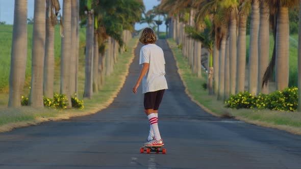 A Teenager with Long Hair Rides a Skateboard Along a Beautiful Road with Palm Trees