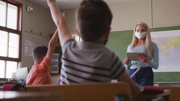 Female teacher wearing face mask pointing towards a boy raising his hands in class at school