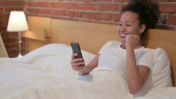 African Woman Celebrating Success on Smartphone in Bed