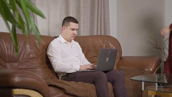 Absorbed Concentrated Caucasian Man Messaging Online on Laptop As Pregnant Woman Entering Room and