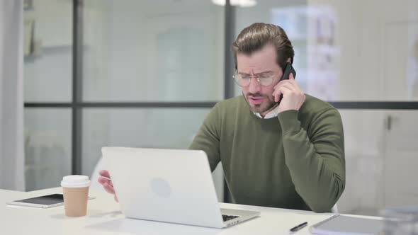 Angry Young Businessman Talking on Smartphone While Using Laptop in Office