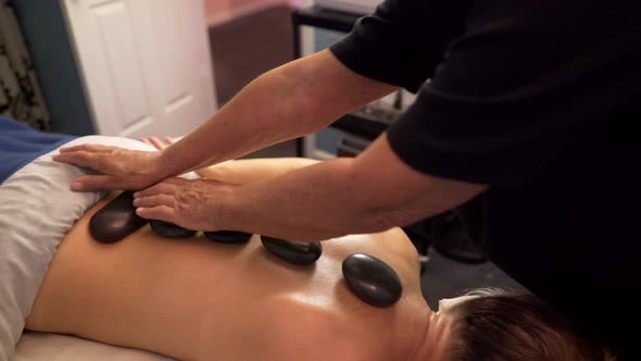 Masseuse or massage therapist uses heated stones on mature, beautiful woman’s back and hands. Steadi